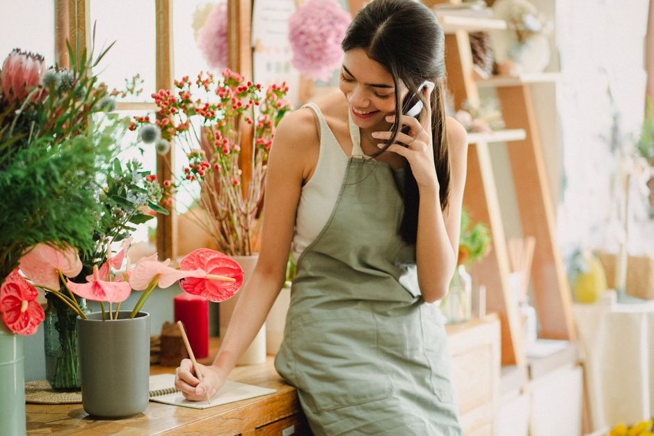 A young woman with long dark hair, dressed in a sleeveless white top and a beige apron, is smiling as she writes on a note pad with her right hand in a flower shop. She is holding a mobile phone to he