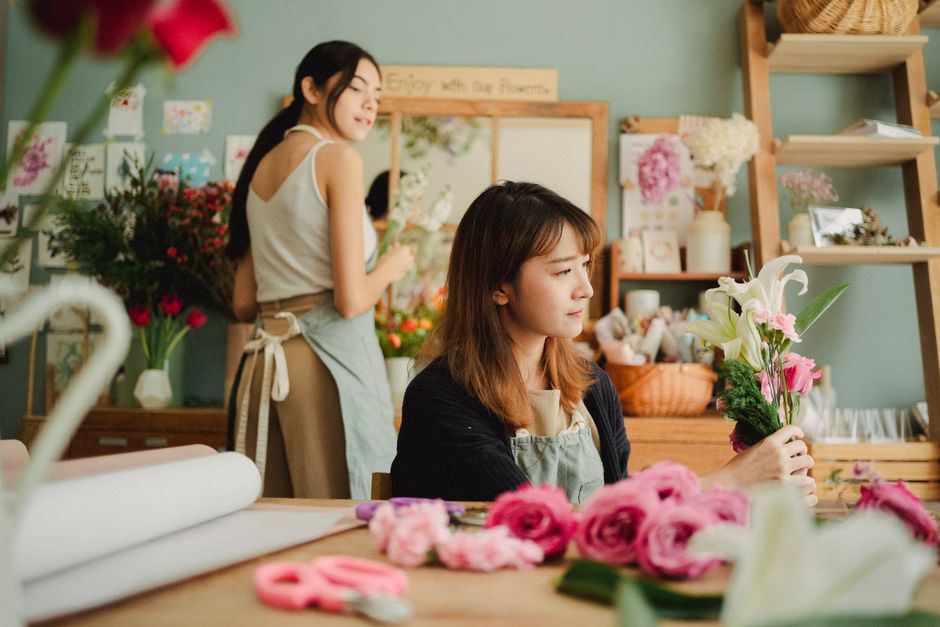 A young woman with shoulder-length brown hair, wearing a light-colored shirt and a grey apron, is seated at a wooden table in a floral workshop, carefully arranging a bouquet of white lilies, pink ros