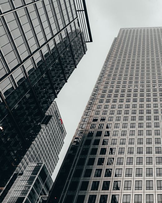 The image captures a low-angle view of modern skyscrapers with glass facades reflecting the overcast sky, emphasizing their height and sleek architectural design in an urban business district. The cen