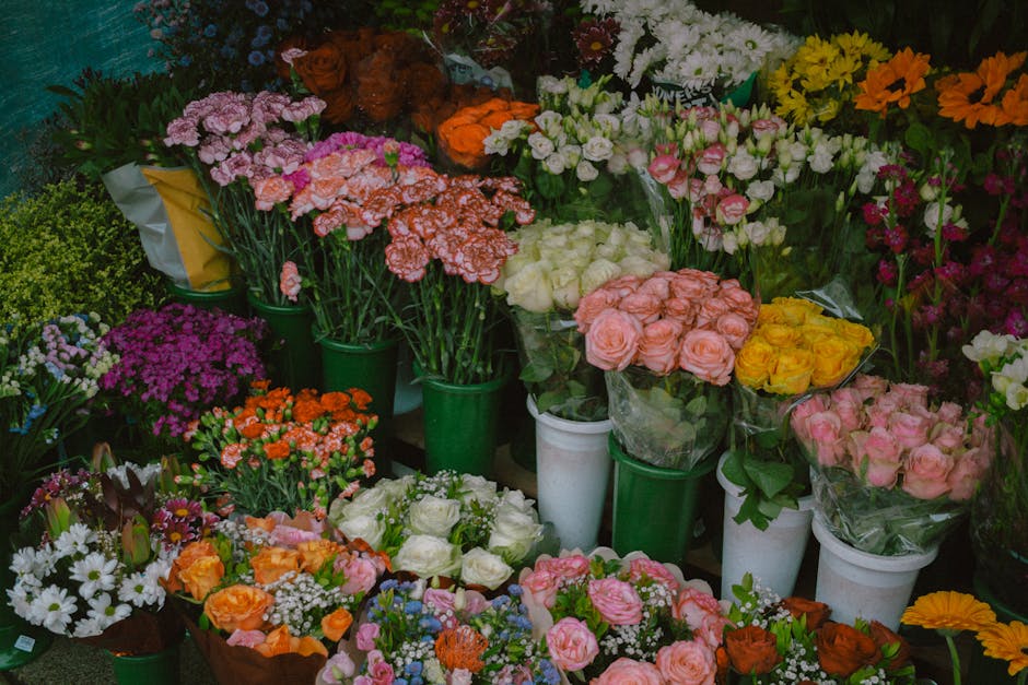 A vibrant display of freshly cut flowers arranged in various floral arrangements and bouquets. The collection includes pink, white, yellow, orange, and purple blooms, such as roses, carnations, daisie