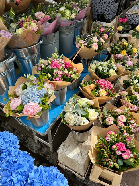 A vibrant display of fresh flower bouquets arranged in metal buckets, wrapped in brown craft paper, and placed on tables at a flower market. The arrangements feature a variety of flowers including pin