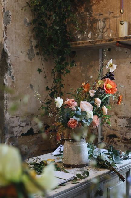 A floral arrangement featuring a mixed bouquet of fresh flowers, including white roses, peach and orange poppies, and various greenery, arranged in a tall, textured ceramic vase placed on a wooden tab