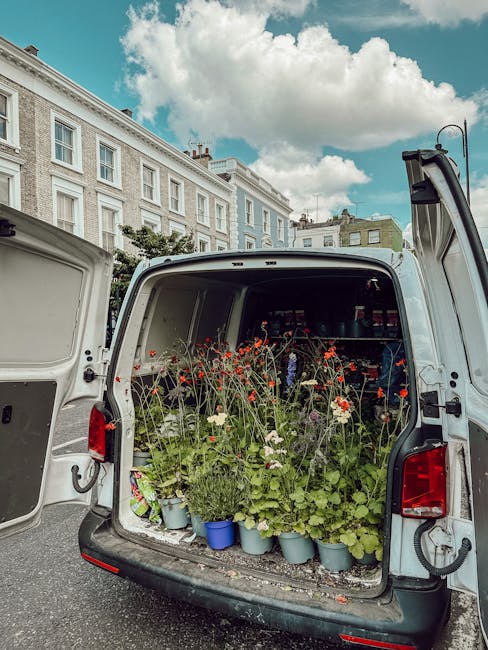 The back of a white delivery van opened to reveal an arrangement of fresh flowers, including red poppies, white daisies, and green foliage in light blue pots and scattered around the vehicle's interio