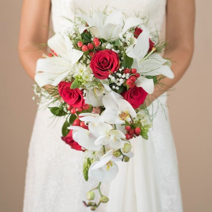 Radiating grace amid the soft, natural light associated with Limehouse's waterside neighborhoods, this bridal bouquet stands as a dazzling centrepiece in a bride's gentle embrace. The medium close-up reveals her arms, elegantly tanned and bare, cradling an arrangement abundant in luxury and style. At the heart of the bouquet, impressive white oriental lilies bloom, their broad petals and vibrant stamens shining above a myriad of supporting blossoms. Richly coloured, full-bodied red roses nestle amongst the white, symbolizing undying love-a motif echoed in Limehouse's storied architecture and riverside romance. Tumbling from the bouquet's centre, Phalaenopsis orchids unfurl in delicate streams, their waxy petals delicately marked with yellow, suggesting a sense of movement as fluid as Limehouse Cut itself. Clusters of hypericum berries and wisps of baby's breath (gypsophila) bring lively pops of red and airy, cloud-like softness amid the more structural greenery. Asparagus fern and feathery fronds, paired with deeper greens, wrap around the base and sides, crafting a sense of depth and natural abundance reminiscent of Ropemaker's Fields. The bride's intricate white gown-hints of lace catching the light-further enhances the image's aura of sophisticated romance. This meticulously designed bouquet, steeped in local character and artistry, would be a perfect touch for Limehouse weddings, conjuring both modern elegance and a sense of timeless East London celebration.