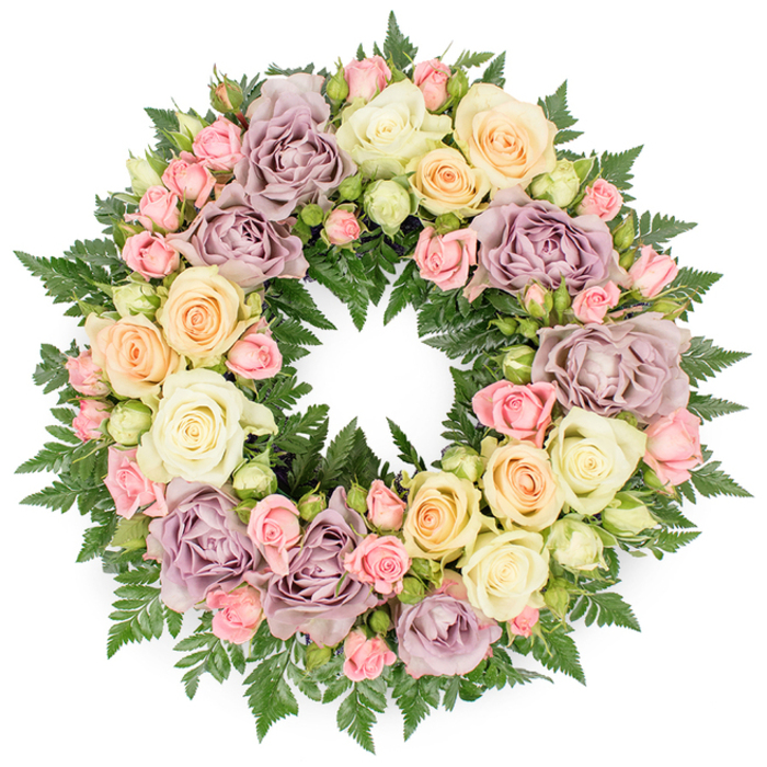 A lush, full circular wreath is shown against a white background, its intricate arrangement of roses and foliage capturing the eye and drawing it around the ring again and again. This is a classic funeral or memorial wreath, but with a softness that feels deeply personal. Large roses in creamy white, pale peach, and dusty lavender or mauve dominate the design, their petals opening wide to reveal layered centres that look almost like carved ivory or folded silk. Between them, clusters of smaller pink spray roses add detail and intimacy, their tiny buds and partially opened blooms suggesting quiet moments and cherished memories. A sprinkle of pale greenish buds hints at freshness and renewal, as if the wreath were assembled just a short walk away from Limehouse Marina before being carried to a nearby service. Dark green fern fronds provide the underlying structure, their finely divided leaves radiating around the circle and pushing gently through the flowers. This rich greenery gives the wreath depth and creates a pleasing contrast with the pastel roses, much like the interplay of glass towers and old brick warehouses that define Limehouse's skyline. The overall lighting is soft and even, revealing the natural texture of the petals and the subtle shine of the foliage without harsh highlights. Emotionally, the piece speaks of tranquillity and honour, a thoughtful floral tribute for a loved one's funeral or memorial gathering in Limehouse or along the riverside. The craftsmanship suggests careful hands and a sympathetic mind, arranging each bloom to express peace, remembrance, and the continuing bond between the departed and those who remain.